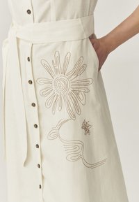 Kermavaale linen-kleid, das mit braunen gestickten Sonnen- und Bienenmotiven verziert ist, vorne mit Knöpfen geschlossen wird und einen gebundenen Taillengürtel hat.