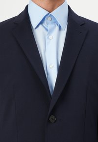 Blazer azul marino con un solo botón negro. Camisa de vestir azul clara debajo con un cuello clásico y botones perlados. Textura de tela suave.