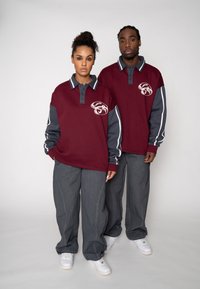 Bordeauxrote Langarm-Poloshirts mit grauen Ärmeln, geknöpftem Kragen und Logo; kombiniert mit weiten grauen Hosen und weißen Turnschuhen, stehend vor einem hellen Hintergrund.