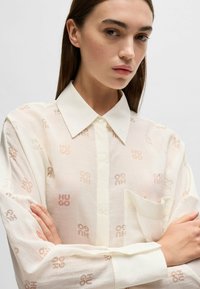Transparante crème blouse met een subtiel herhaald "HUGO" logo in lichtroze. Hij heeft een puntige kraag en een borstzakje.
