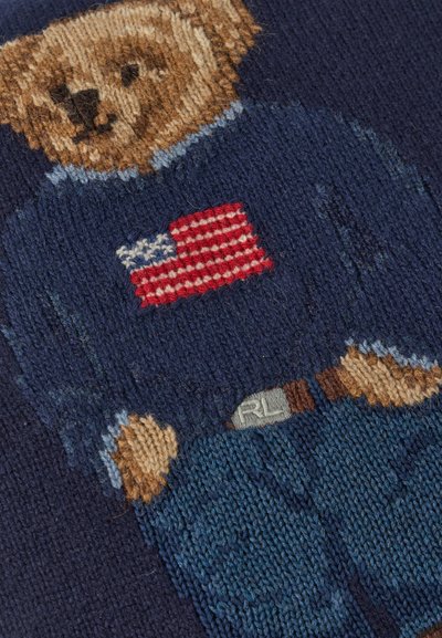 Πλεκτή αρκουδίτσα που φοράει μπλε πουλόβερ με αμερικανική σημαία και μπλε παντελόνι με ζώνη σε σκούρο μπλε φόντο.