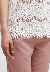 Top in pizzo bianco con bordo scallop e dettagli in frange, sovrapposto a pantaloni in satin rosa chiaro, che mostrano una texture liscia e un design raffinato.