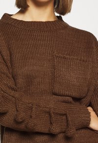 Pull en tricot marron avec un col rond, un tissu texturé, une poche avant et des accents en pompons le long des manches.