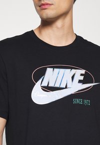Schwarzes Baumwoll-T-Shirt mit einem großen, schimmernden Nike-Logo und dem Text "Champion of Sport" sowie "Since 1972" in kontrastierenden Farben.