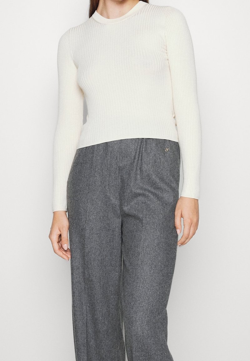 Personne portant un haut blanc côtelé ajusté à manches longues et un pantalon large en laine grise taille haute, debout devant un fond uni.