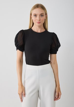 Black puff-sleeve top in smooth fabric, featuring sheer, textured puff sleeves. Paired with high-waisted, white tailored pants.