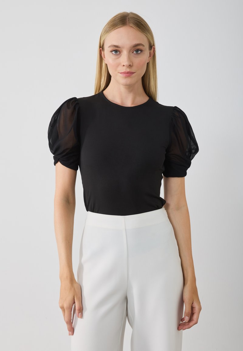 Melns puff-sleeve tops gludā audumā, ar caurspīdīgām, strukturētām puff piedurknēm. Apvienots ar augsta vidukļa, baltām pielāgotām biksēm.