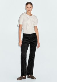 Maglia a maglia beige chiaro con dettaglio annodato allo scollatore; abbinata a pantaloni neri a gamba dritta realizzati in tessuto testurizzato e scarpe flat marroni.