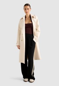 Donna che indossa un trench coat crema sopra un top a collo halter scuro, pantaloni della tuta neri con strisce bianche e scarpe da ginnastica beige, in piedi di fronte a uno sfondo semplice.