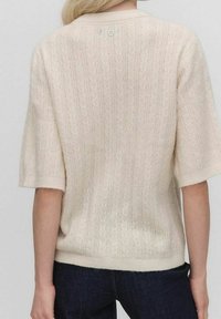 Krémový pletený svetr s texturovaným vzorem, krátkými rukávy a kulatým výstřihem. Obsahuje detail knoflíku na zadní straně.