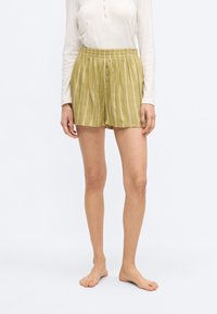 Γυναίκα που φοράει ανοιχτό μπεζ μακρυμάνικη μπλούζα και χαλαρά ανοιχτοπράσινα σορτς με λευκές και ροζ κατακόρυφες ρίγες, σκέτη, σε λευκό φόντο.