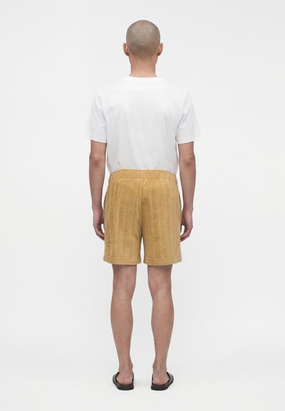 Homme debout de dos portant un t-shirt blanc, un short beige texturé et des sandales noires sur un fond blanc uni.