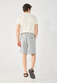 Chemise beige clair à manches courtes associée à un short gris avec une taille élastique et deux poches arrière. Des sandales noires complètent la tenue.