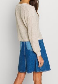 Woman wearing a beige knitted sweater and a two-tone blue denim skirt, standing sideways against a plain light background.