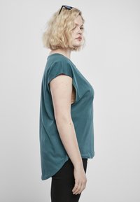Teal bomullströja med korta, rullade ärmar och en lös passform. Har en sidoklippdetalj och en rundad nederkant, bärs över svarta byxor.