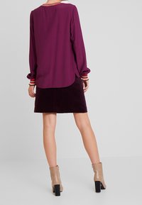 Blusa viola a maniche lunghe con polsini a righe, abbinata a una gonna in velluto a coste di un viola scuro. La modella indossa stivaletti beige con tacco largo.