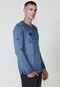 Rövid hajú férfi, aki kék, hosszú ujjas grafikás pólót és fekete nadrágot visel, az egyik kezét a zsebébe dugva, enyhe szögben, előre néz.
