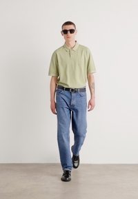 Polo shirt vert clair avec logo, jeans bleus, chaussures noires et lunettes de soleil. La tenue présente une coupe décontractée et un design casual.