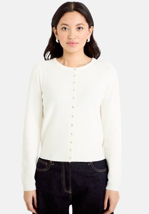 Vrouw draagt een witte gebreide cardigan met parelachtige knopen en donkere denim jeans, staand tegen een lichte achtergrond.