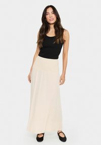 Fekete tank top, amit egy krémszínű, smocked derékkal ellátott maxi szoknyával párosítottak. A szoknya lágyan hullámzó, sima textúrájú. Fekete, pántos cipők.