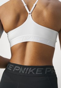 White sports bra with thin straps and embossed "Nike" logo on the band. Worn with high-waisted black leggings featuring a grey "Nike" print.