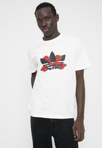 Λευκό κοντομάνικο μπλουζάκι με μαύρο λογότυπο τριών φύλλων, διακοσμημένο με κόκκινα τριαντάφυλλα και πράσινα φύλλα· κατασκευασμένο από απαλό βαμβακερό ύφασμα.