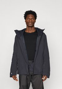 Homem a usar uma jaqueta com capuz escura, aberta, sobre uma camisa preta e calças escuras, de pé contra um fundo claro e simples.