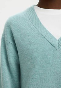 Soft, light blue knitted sweater with a V-neck, featuring a textured pattern along the collar and relaxed fit. Layered over a white top.