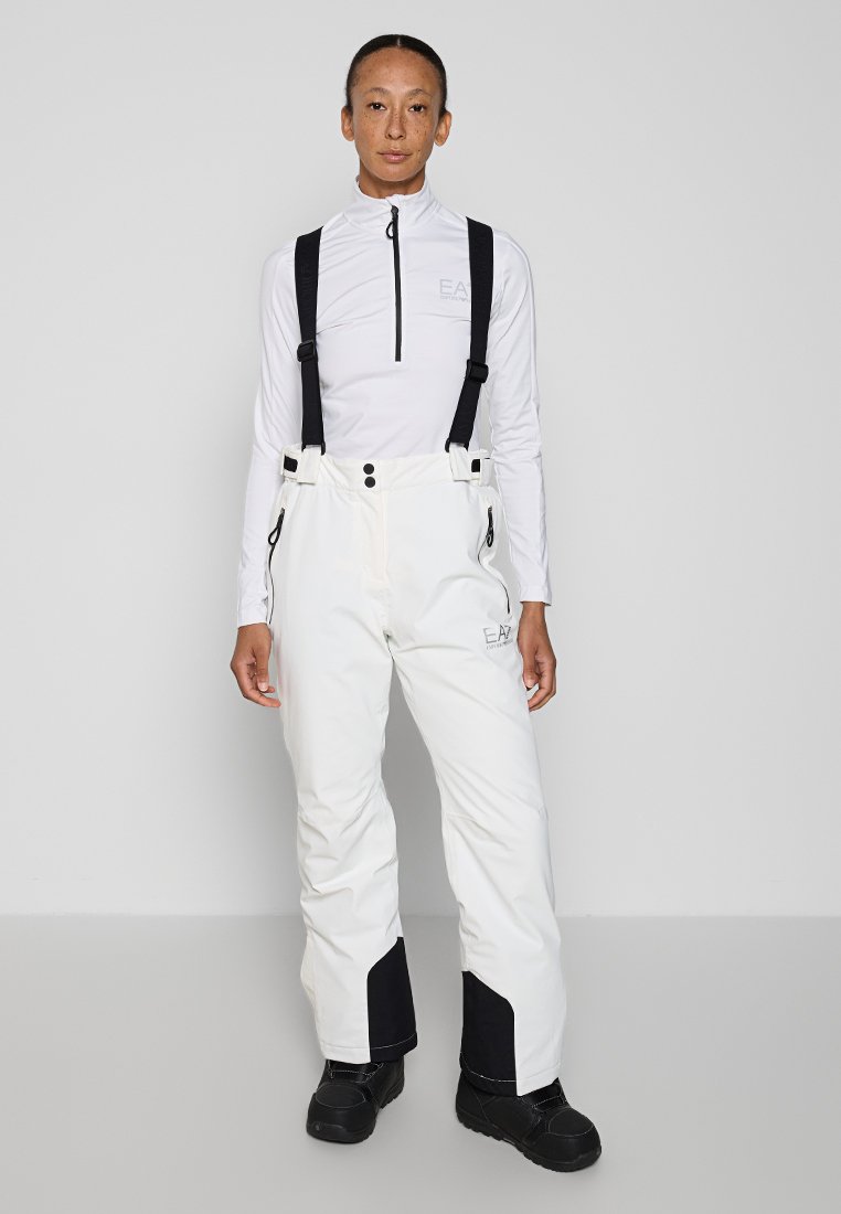 Pantaloni da sci bianchi con bretelle nere regolabili, tasche con zip e dettagli neri alle caviglie. Abbinati a un top bianco a maniche lunghe.