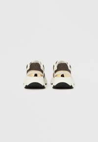 Lenkkitossut, joissa on valkoinen pohja, ruskea kuvioitu yläosa ja kultaiset kantakoristeet. Varustettu pehmustetulla kauluksella ja kuvioidulla verkkopaneelilla.