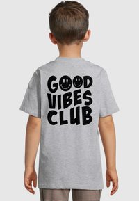 Šedé tričko s krátkým rukávem s výrazným černým textem "GOOD VIBES CLUB" a dvěma usmívajícími se tvářemi na zadní straně. Měkký bavlněný materiál.