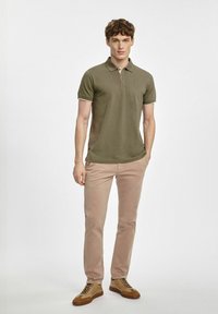 Oliwkowa koszulka polo z kołnierzykiem, z kontrastowym wykończeniem przy rękawach, zestawiona z jasnobezowymi spodniami slim-fit oraz brązowymi trampkami z sznurowadłami.