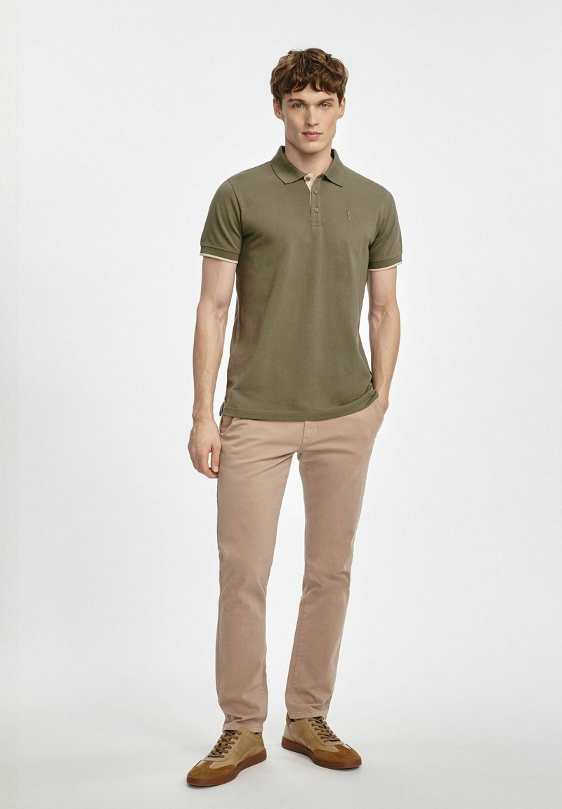 Oliwkowa koszulka polo z kołnierzykiem, z kontrastowym wykończeniem przy rękawach, zestawiona z jasnobezowymi spodniami slim-fit oraz brązowymi trampkami z sznurowadłami.