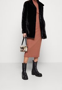 Abrigo de piel sintética negra, vestido midi ajustado marrón, bolso estampado y botas de tacón grueso y negro. Textura suave, diseño elegante y detalles modernos.