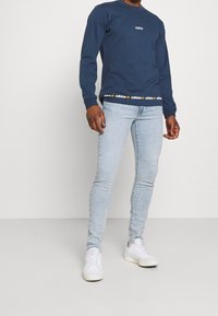 Námořnická modrá košile s dlouhým rukávem a kulatým výstřihem, se znakem adidas a páskem s opakujícím se vzorem loga, spárovaná s světle modrými džíny.