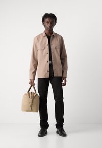 Lys beige knap-ophængt jakke med to brystlommer, sort T-shirt, sorte bukser og en tan duffelbag med lynlås og læderdetaljer.