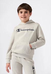 Hymyilevä poika, joka on pukeutunut vaaleanharmaaseen Champion-huppariin ja samankaltaisiin verkkohousuihin, joissa on logoja. Hän seisoo yksi käsi hupparin Taskussa.