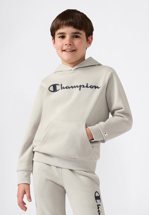 Un garçon souriant portant un sweat à capuche Champion gris clair et un pantalon de survêtement assorti avec des logos, se tenant avec une main dans la poche du sweat.