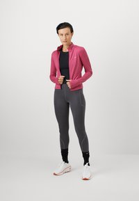 Nainen vaaleanpunaisessa vetoketjullisessa takissa, mustassa paidassa, harmaissa leggingsseissä, mustissa säärystimissä ja valkoisissa juoksukengissä seisoo yksinkertaista taustaa vasten.