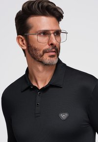 Sort langærmet polo-shirt med struktureret krave, tre-knaps knaplukning og en lille logo-badge på venstre bryst. Gennemsigtige briller.