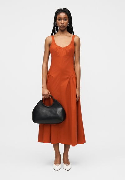 Γυναίκα με πλεγμένα μαλλιά, φορούσα φορέμα μέσης μήκους σε σκουρό πορτοκαλί απόχρωση χωρίς μανίκια, κρατώντας μια μαύρη τσάντα χειρός και φορώντας αιχμηρά λευκά παπούτσια.