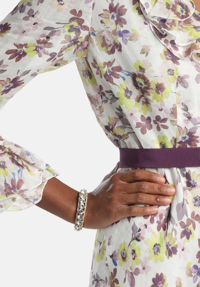 Roka na boku, oblečena v cvetlično prosojno bluzo z nabranimi manšetami, vijoličnim pasom in bleščečo kristalno zapestnico na rjavi koži.