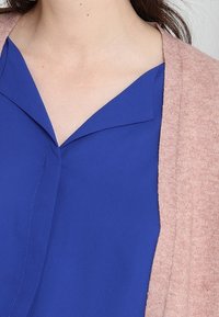 Blå blus med V-ringning och korta ärmar, lagd under en mjuk rosa cardigan. Tyget verkar slätt med en subtil lyster.