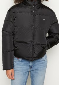 Doudoune noire avec un col haut, dotée d'une fermeture à boutons et d'un design matelassé. Associée à un jean en denim bleu clair.