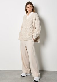 Beige grob gestrickter Pullover mit Rautenmuster, kombiniert mit lockeren cremefarbenen Hosen und weißen Sneakers. Die Textur wirkt dick und gemütlich.