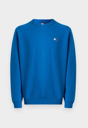 Sininen huppari, joka on valmistettu pehmeästä kankaasta ja jossa on ribattu pyöreä kaula-aukko ja hihat. Vasemmassa rintataskussa on pieni logopatch.