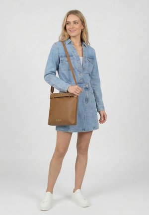 Kvinde iført en denimkjole og hvide sneakers, der bærer en brun læder crossbody taske med bælte-detalje.