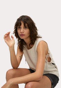 Top sin mangas a rayas en tejido ligero, con lazos laterales y un corte holgado. La modelo lleva pantalones cortos oscuros, con un enfoque en el escote y la textura.