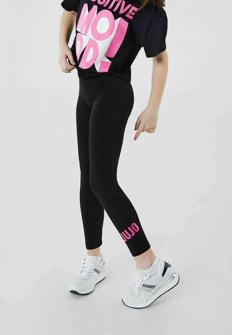 Černé legíny s růžovým textem, doplněné černým tričkem s krátkým rukávem, na kterém je velký růžový a bílý grafický text. Bílé tenisky se stříbrnými detaily.