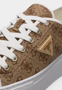 Κοντινό πλάνο καφέ αθλητικού παπουτσιού Guess με λευκά κορδόνια, υφασμάτινο με σχέδια, χρυσά θηλύκια και ανάγλυφο τρίγωνο λογότυπο Guess στο πλάι.